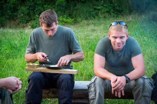 Zwei Personen sitzen auf einer Holzbank, eine schneidet mit einem Messer