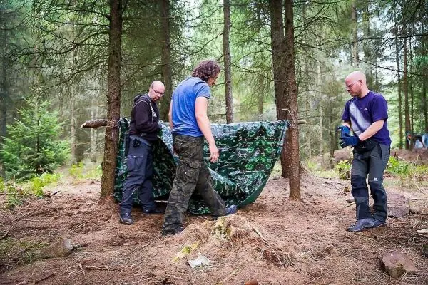 Drei Personen bauen eine Tarp-Notunterkunft zwischen Bäumen auf
