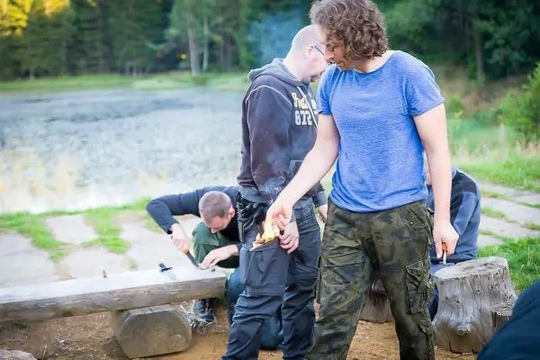 Feuerstahl entzündet Zunder am Boden, während andere an einer Holzstruktur arbeiten
