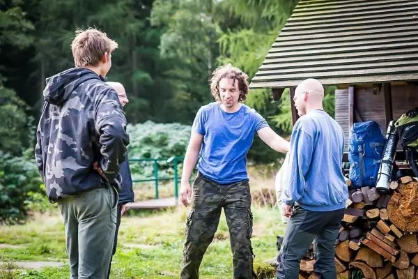 Gruppe diskutiert vor einem Holzstapel und Rucksäcken unter einem Dach