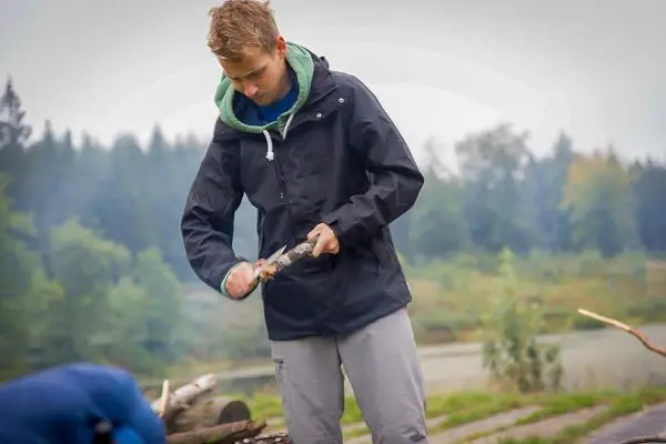 Junge schnitzt mit einem Messer an einem Holzstück am Ufer eines Gewässers