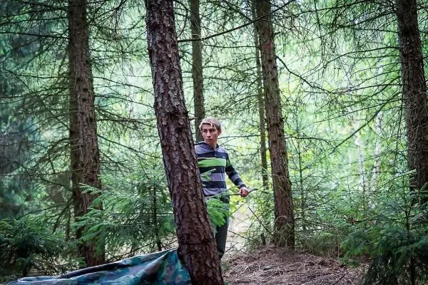 Junge steht zwischen Bäumen in einem Wald, Tarp liegt am Boden