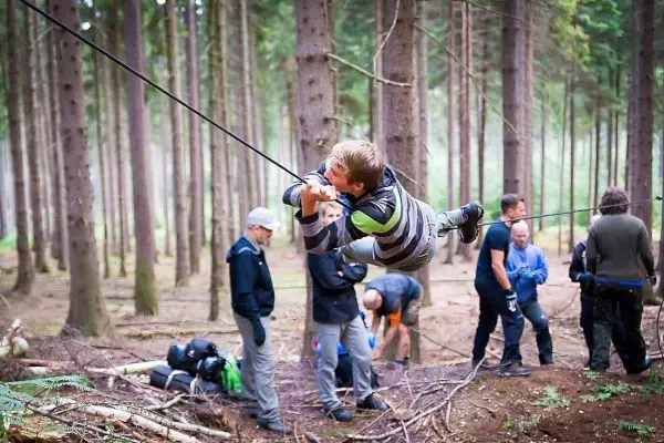 Junge überquert selbstgebaute Seilbrücke zwischen Bäumen, Gruppe beobachtet