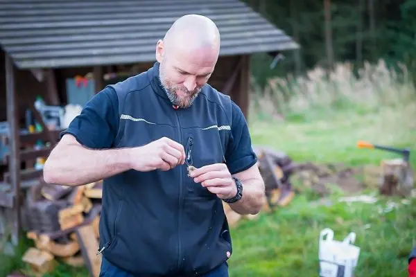 Mann bearbeitet ein kleines Werkzeug in der Hand, im Hintergrund Holzstapel
