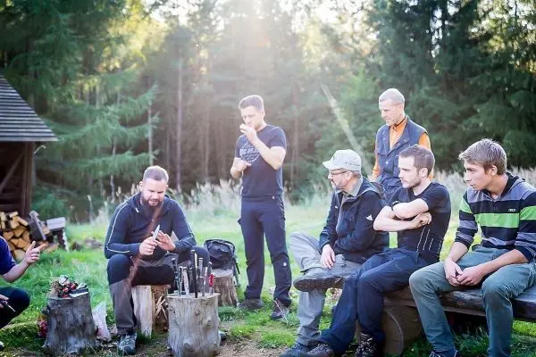 Mann bearbeitet Messer auf Holzstamm, Gruppe beobachtet aufmerksam