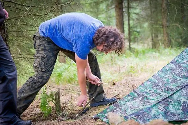 Mann befestigt eine Tarp-Notunterkunft mit einem Stock im Wald
