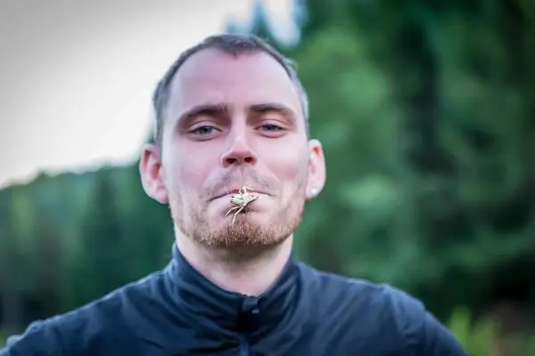 Mann hält eine Heuschrecke im Mund und lächelt in die Kamera