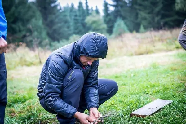 Mann kniet im Gras und bearbeitet ein Holzstück mit einem Messer