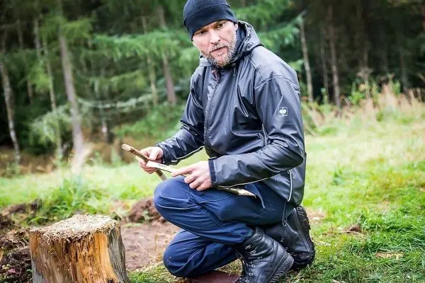 Mann kniet neben einem Baumstumpf und bearbeitet einen Stock mit einem Messer