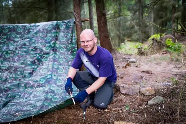 Mann kniet neben einer Tarp-Notunterkunft und hält ein Messer in der Hand
