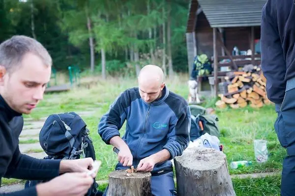 Mann schnitzt mit einem Messer an einem Holzblock, Rucksäcke im Hintergrund