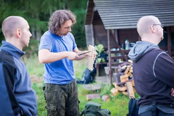 Mann schnitzt mit einem Messer an einem Holzstück, während andere zuschauen