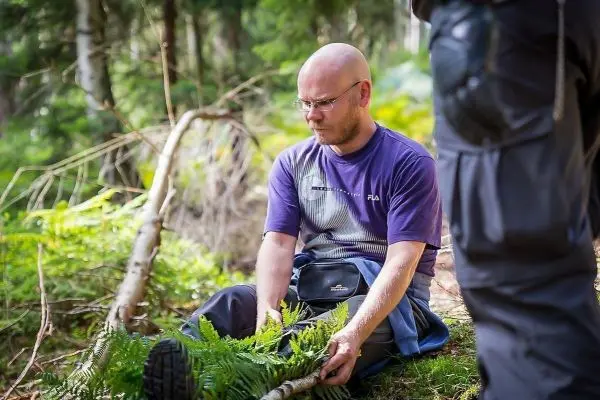 Mann sitzt im Wald und bearbeitet einen Ast mit Farnen