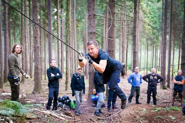 Mann überquert selbstgebaute Seilbrücke im Wald, Zuschauer beobachten