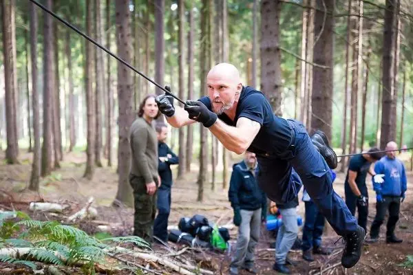 Mann überquert selbstgebaute Seilbrücke im Wald, Zuschauer im Hintergrund