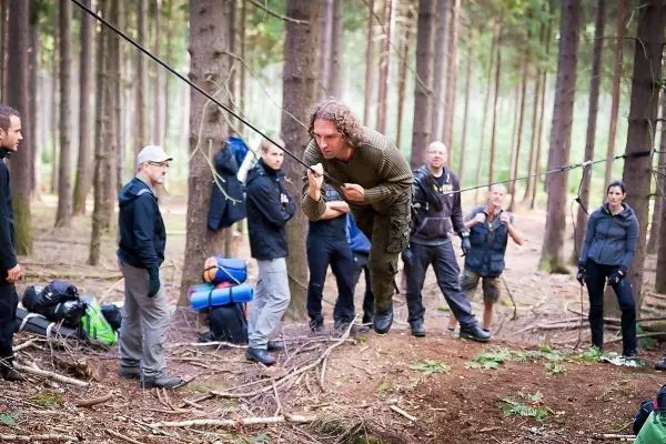 Mann überquert selbstgebaute Seilbrücke zwischen Bäumen, Zuschauer beobachten