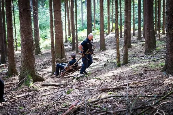 Mann zieht improvisierte Trage durch den Wald mit einem weiteren Mann darauf