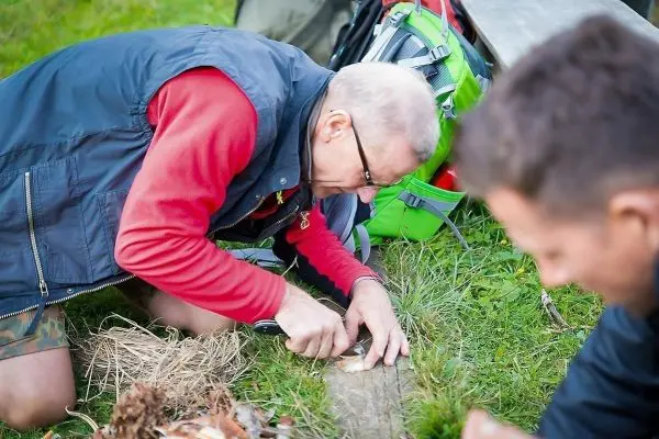 Messer schneidet Holzstück auf einer Wiese, Zunder liegt daneben