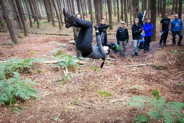 Person überquert selbstgebaute Seilbrücke zwischen Bäumen, Zuschauer beobachten