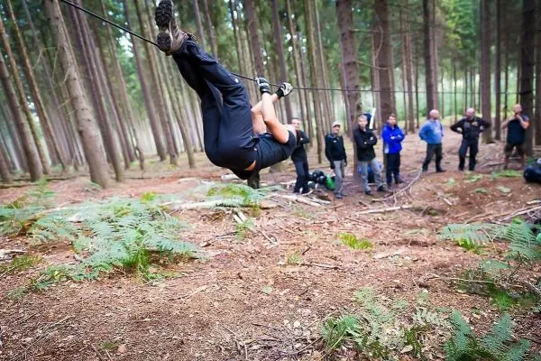 Person überquert selbstgebaute Seilbrücke zwischen Bäumen, Zuschauer beobachten
