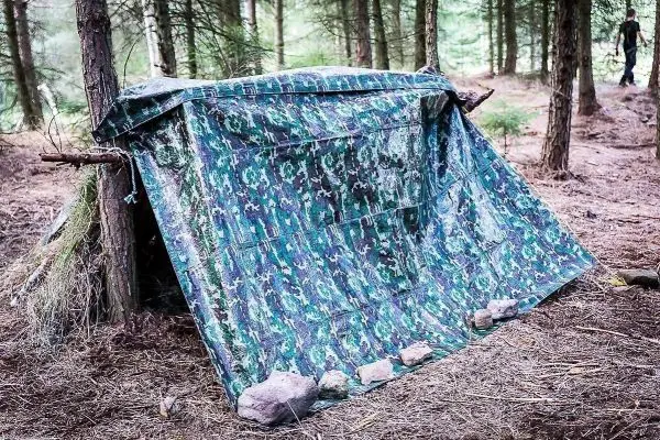 Tarp-Notunterkunft aus Camouflage-Material, mit Steinen beschwert, im Wald aufgebaut