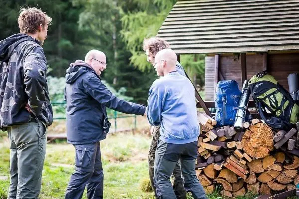 Vier Personen stehen in einer Waldumgebung neben einem Holzstapel und Rucksäcken