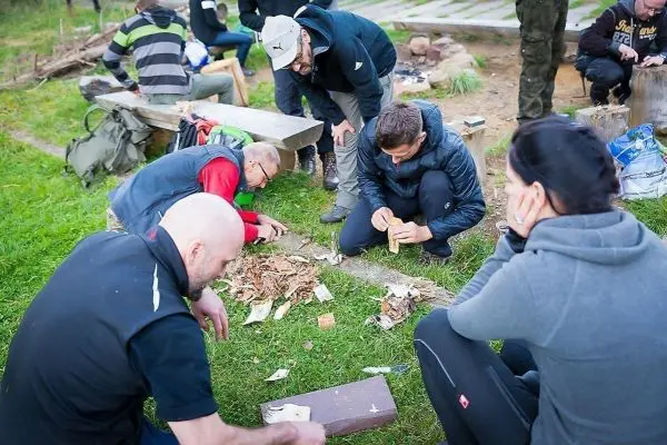 Zunder wird auf einem Holzstück vorbereitet, während Teilnehmer umher sitzen