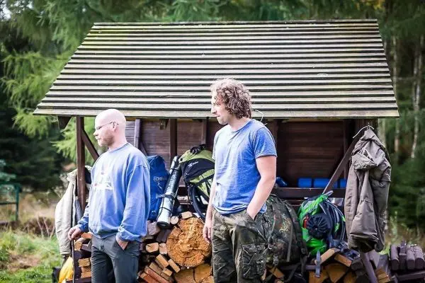 Zwei Männer stehen neben einem Holzstapel und einem Unterstand mit Rucksäcken