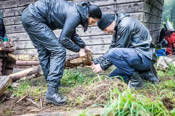 Zwei Personen bearbeiten einen Holzstock auf dem Boden neben einer Hütte