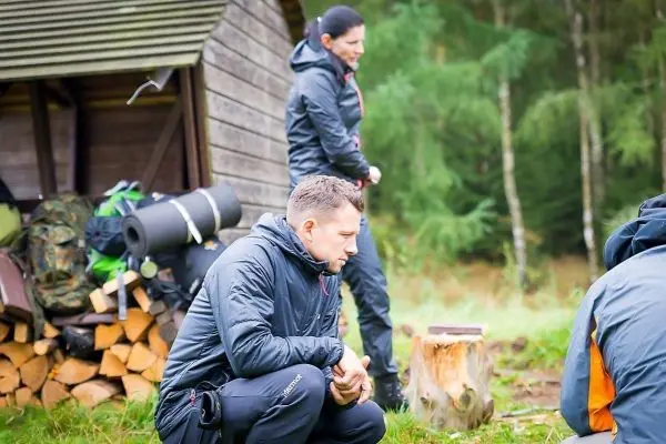 Zwei Personen in Outdoor-Bekleidung stehen und knien vor einem Holzstapel