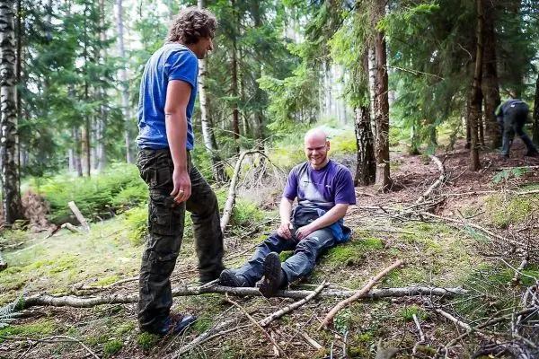 Zwei Personen sitzen im Wald auf dem Boden, eine Person hilft der anderen