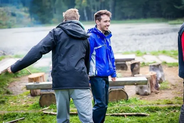 Zwei Personen stehen an einem Lagerplatz mit Holzstämmen und Blick auf einen Gewässerbereich