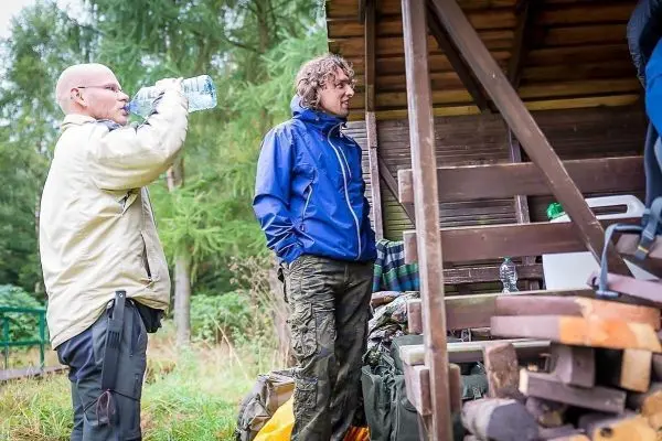 Zwei Personen stehen an einer Holzunterkunft, einer trinkt Wasser aus einer Flasche