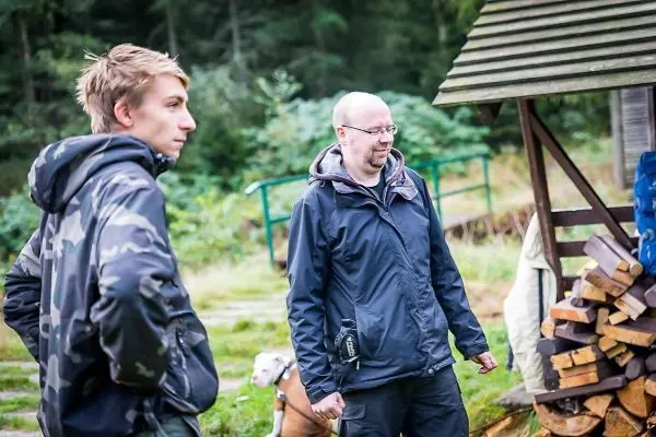 Zwei Personen stehen neben einem Holzstapel in einer Waldumgebung