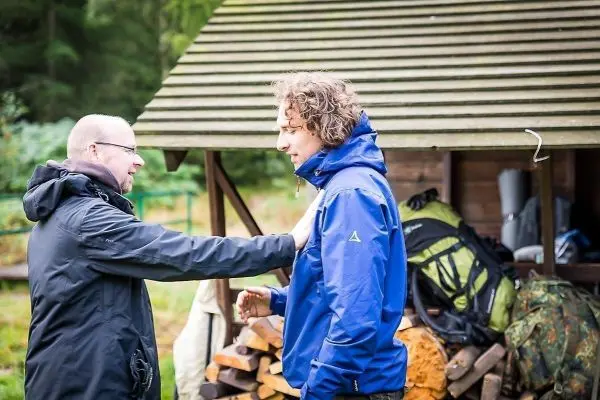 Zwei Personen stehen vor einer Holzunterkunft und interagieren miteinander