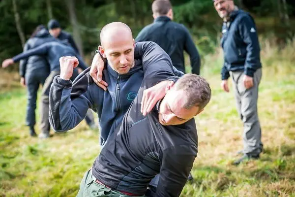 Zwei Personen üben einen Nahkampfgriff auf einer Wiese, weitere Teilnehmer im Hintergrund