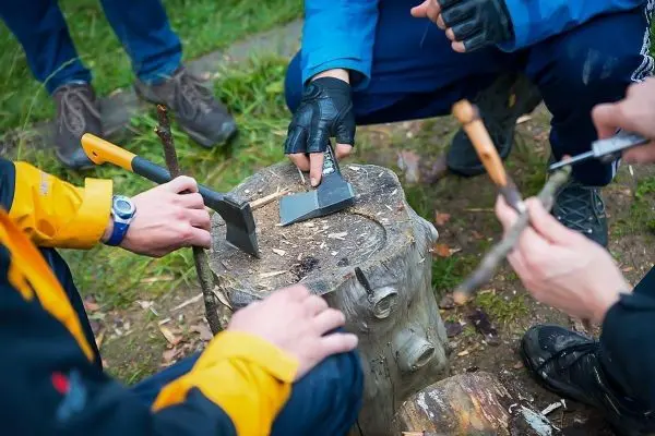 Holzstück wird mit Axt bearbeitet, während mehrere Hände Werkzeuge halten