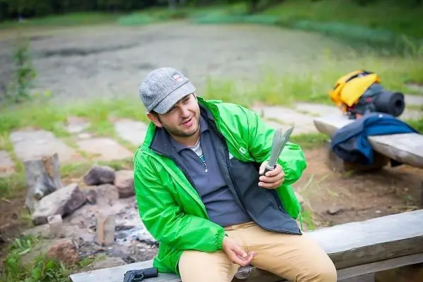 Mann hält ein Messer in der Hand, sitzt an einer Holzbank neben einem kleinen Feuer