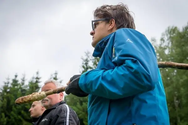 Mann hält einen Stock in der Hand, im Hintergrund weitere Personen und Bäume