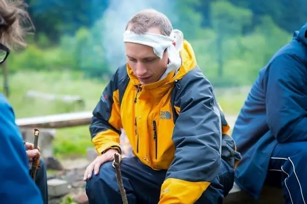 Mann in gelber Jacke sitzt neben einem kleinen Feuer und bearbeitet Holzstücke