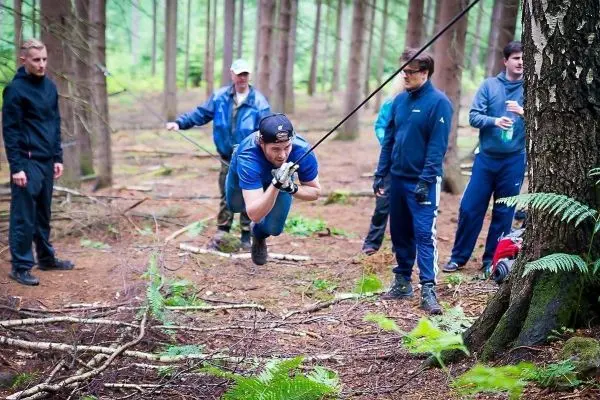 Mann überquert selbstgebaute Seilbrücke zwischen Bäumen, umgeben von Zuschauern