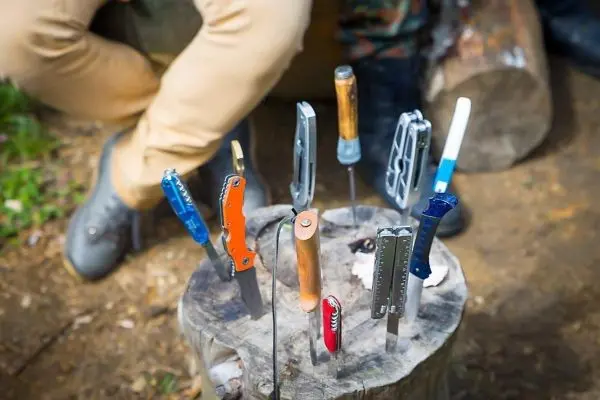 Mehrere Messer stecken in einem Baumstumpf, Person sitzt daneben