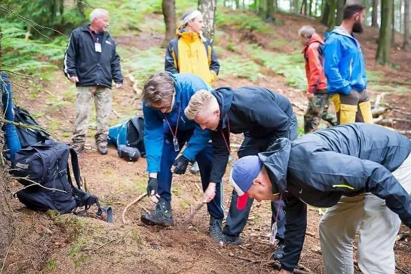 Teilnehmer graben im Waldboden, während Rucksäcke im Hintergrund liegen