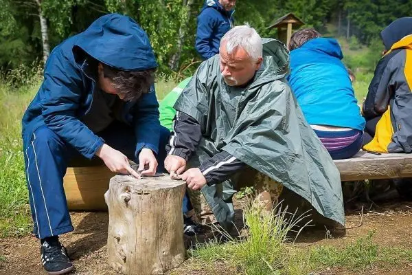 Zwei Personen bearbeiten einen Holzblock mit einem Messer auf einer Holzbank