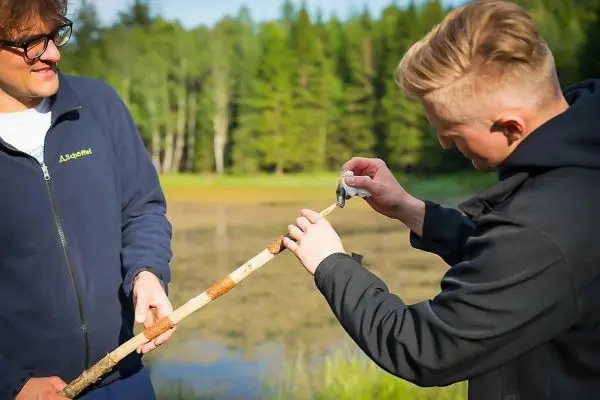 Zwei Personen bearbeiten einen Stock mit einem Messer am Ufer eines Gewässers