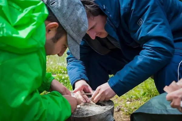 Zwei Personen bearbeiten Holz auf einem Baumstumpf beim Überlebenstraining