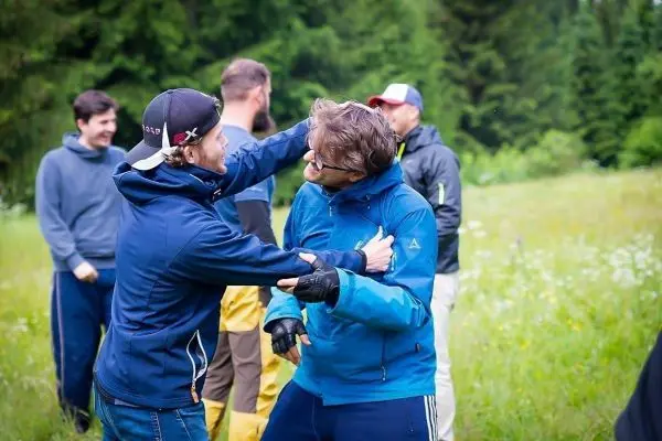 Zwei Personen umarmen sich spielerisch auf einer Wiese, im Hintergrund weitere Teilnehmer