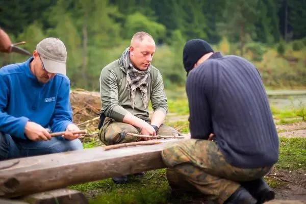 Drei Personen bearbeiten Holzstücke auf einem Baumstamm am Ufer eines Gewässers