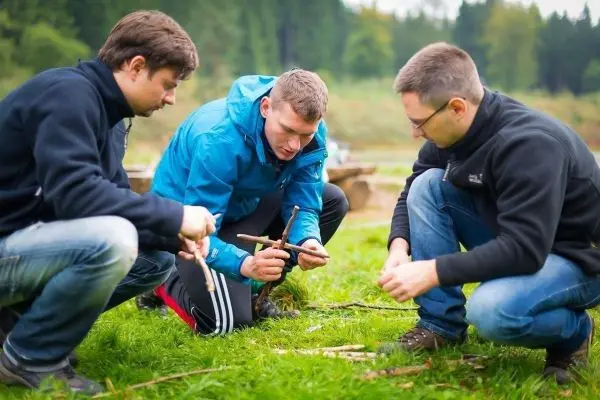 Drei Personen knien auf dem Boden und bearbeiten Holzstücke mit einem Messer