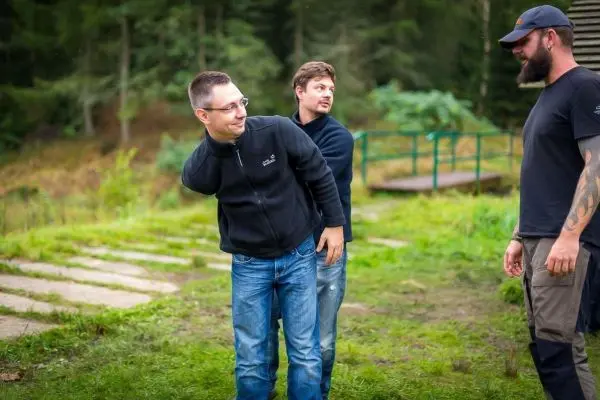 Drei Personen stehen auf einer Wiese, eine Person beugt sich nach vorne